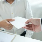 Zwei Personen überreichen einen Umschlag, passend zum Thema Mitarbeitergeschenke steuerlich absetzbar und deren korrekte betriebliche Dokumentation.